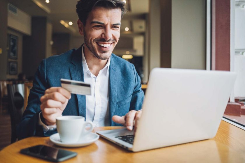 A businessman smiles at his laptop, with a payment card in his hand.