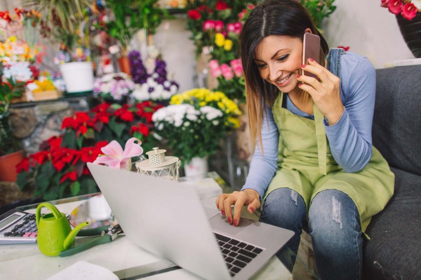 A florist making a call whilst looking at her laptop in a flower shop.