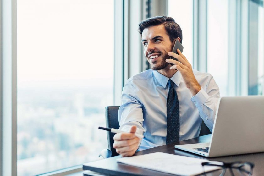 A man on the phone, sitting in an office next to a large window.